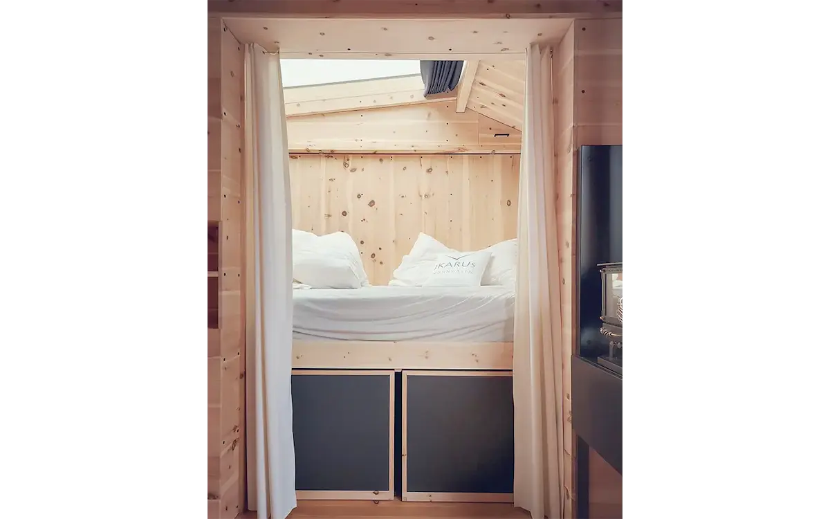 Innenansicht eines Ikarus-Wohnwagens mit Blick auf das erhöhte Bett, Holzverkleidung aus Fichtenholz, weissen Vorhängen und Dachfenster