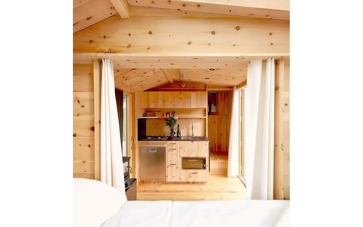 Blick vom Schlafbereich des Ikarus-Wohnwagens durch weisse Vorhänge auf die Küchenzeile mit Edelstahlkühlschrank und Holzausstattung