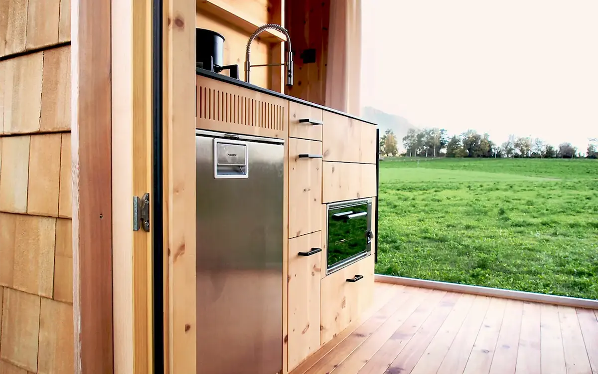 Detailansicht der Küchenzeile des Ikarus-Wohnwagens mit geöffneter Holztür, Blick auf grüne Wiese und Berglandschaft im Hintergrund